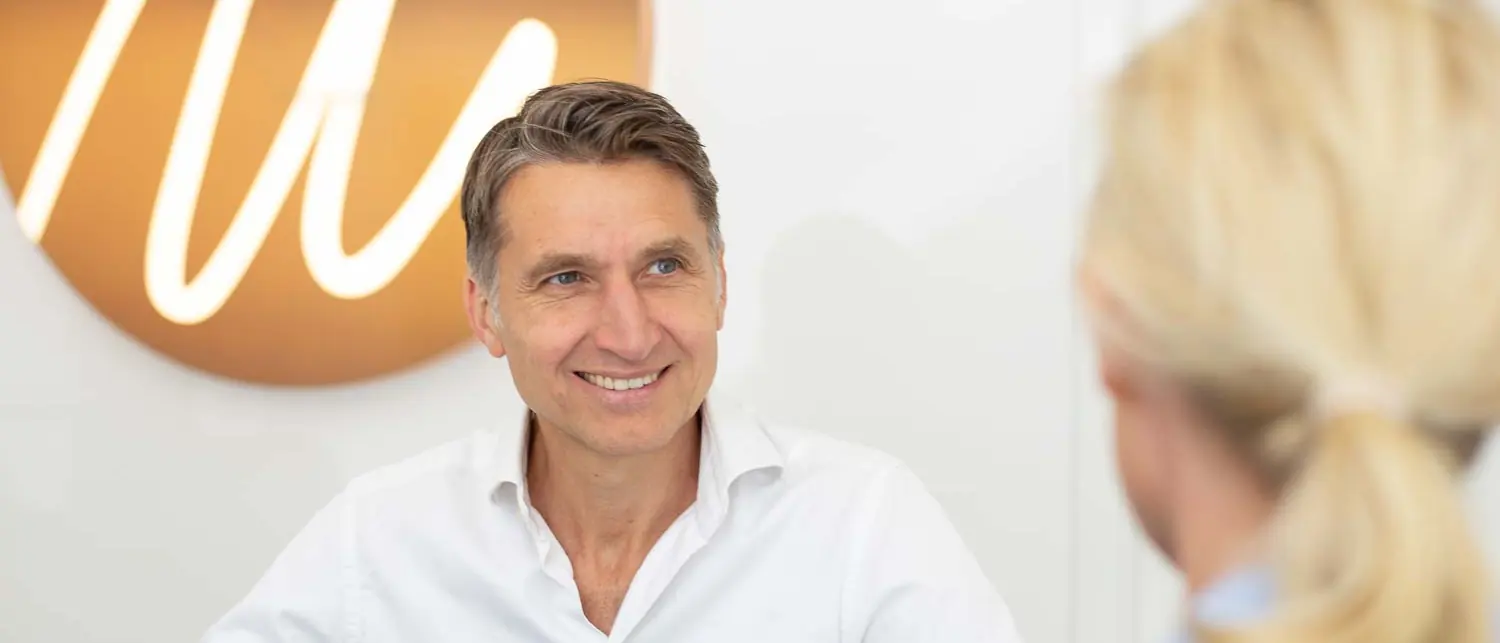 Dr. med. Edouard Manassa im Beratungsgespräch, im Hintergrund das beleuchtete Signet der Praxis